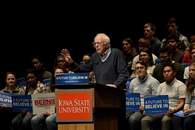 Bernie Sanders at podium  in front of audience holding signs. Flickr photo by Alex Hansen