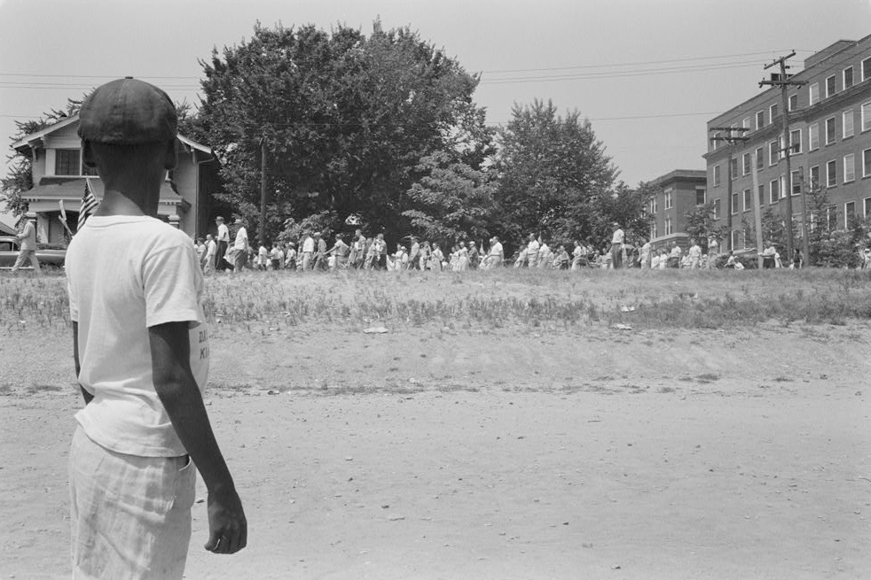 A young African American boy watching a group of people, some carrying American flags, march past to protest the admission of the Little Rock Nine to Central High School
