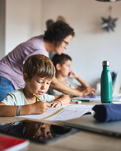 two young learners at table with adult woman helping the boy in the background 