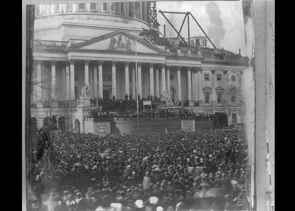 Salted paper print capturing distant view of President Abraham Lincoln at the U.S. Capitol, Washington, D.C. in 1861. He is standing under a wood canopy in front of a crowd.