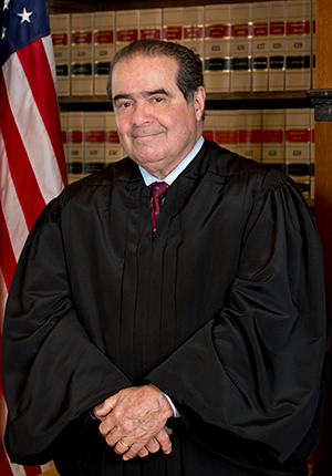 Antonin Scalia, three-quarters portrait, standing in front of U.S. flag and bookcase wearing judicial robes.