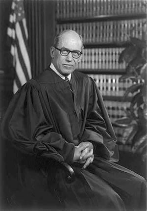 Justice Byron R. White, three-quarters portrait, seated in front of U.S. flag and bookshelf, wearing judicial robes, by Robert S. Oakes, photographer.