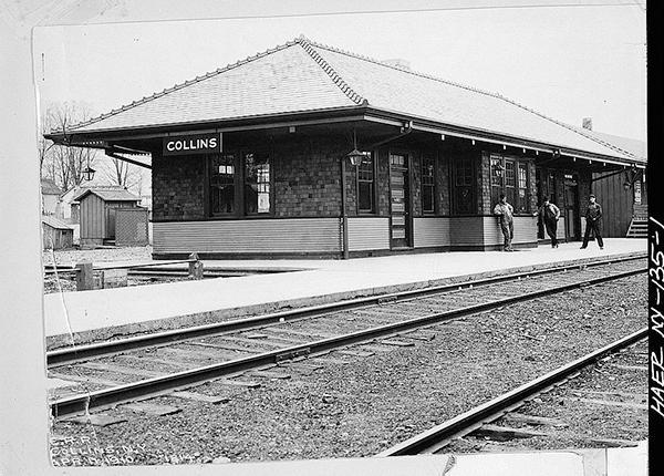 Railway station, with concrete platform, and rail tracks in the foreground. Three men standing on the platform. A sign hanging from the side of the station reads, 'COLLINS'. Photograph taken by Erie Railway Company photographer.