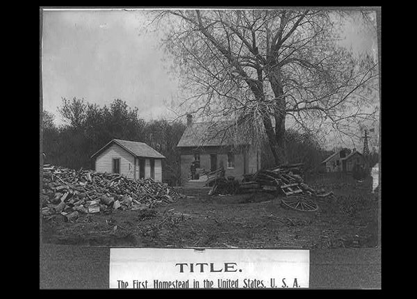 Black and white photograph of man standing on ramp in front of Daniel Freeman homestead in Gage County, Nebraska, the first homestead claim under the 1862 Homestead Act; large piles of wood in yard.