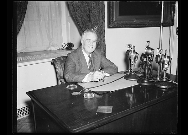 Franklin Delano Roosevelt sitting at table with eyeglasses in one hand. On the table are several broadcast microphones labeled with various radio channels. Harris and Ewing, photographers, 1938.