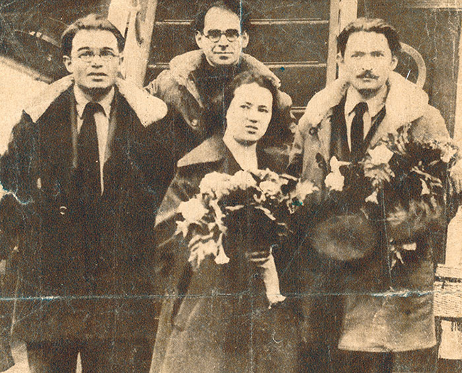 Weathered sepia photo of four adults, three men and one woman, dressed in winter coats. The woman and man on the right are holding flowers.