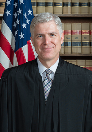 Neil Gorsuch, head-and-shoulder portrait wearing judicial robes, standing in front of U.S. flag and bookcase.