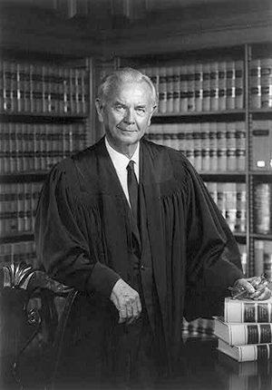 William J. Brennan, Jr. three-quarters portrait, standing in front of bookcase with stack of books to the side, arm resting on chair, wearing judicial robes, by Robert S. Oakes, photographer.