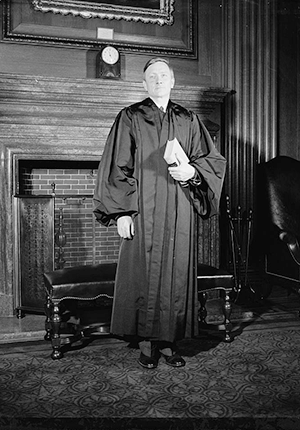 Justice William O. Douglas, standing in front of wooden fireplace wearing judicial robes and holding a book. Photograph by Harris and Ewing, 1939.
