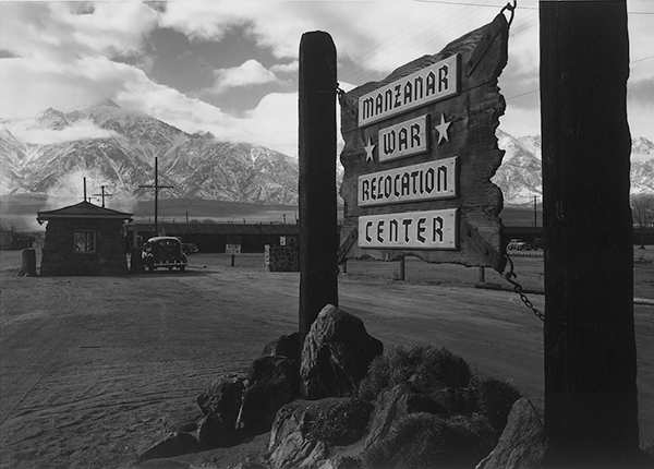 Wooden sign at entrance to the Manzanar War Internment Camp with car and mountains in the background.