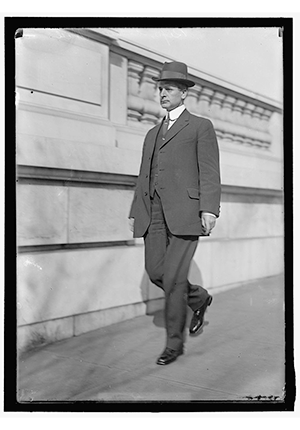 Representative Cordell Hull walking down sidewalk next to building wearing suit and hat. Harris and Ewing, photographers., 1914.
