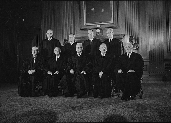 Nine men, five seated and four standing, all in judicial robes, in front of mantel with clock, by Warren K. Leffler, photographer