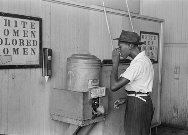 Black man with hat drinking from cup at a water fountain labeled with a sign saying 'colored'. There are two other signs on the wall referring to 'white men, colored men' and 'white women, colored women.'